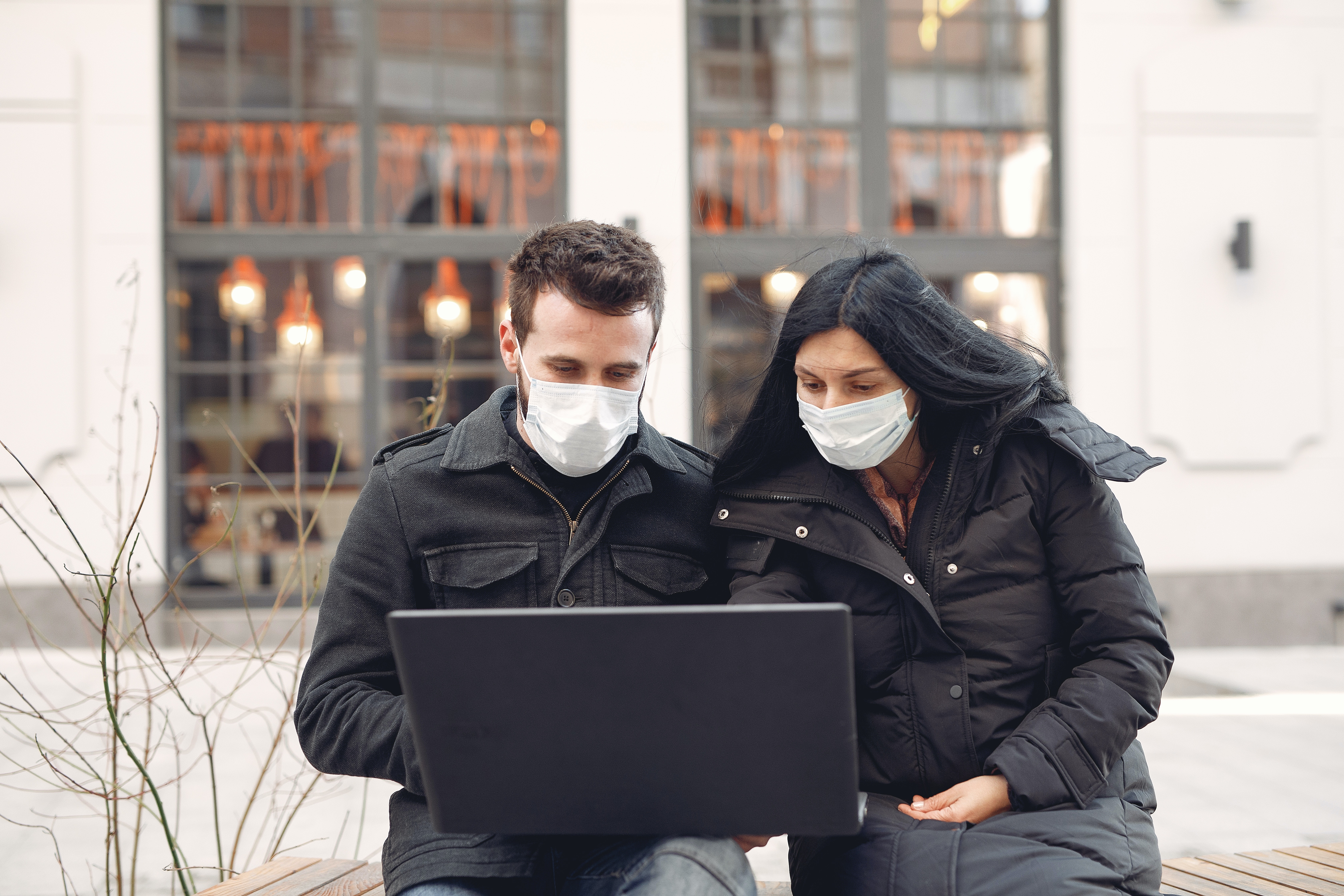 Two people wearing face masks, standing in front of a building and looking at a laptop